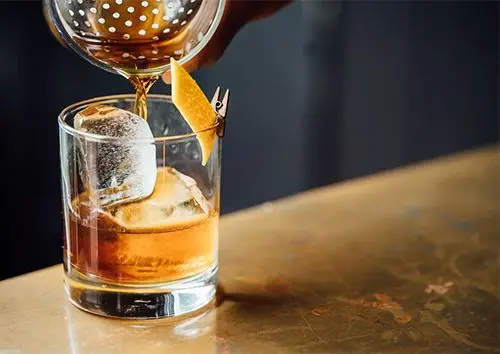 Craft cocktail with an orange twist being strained over a large ice cube.
