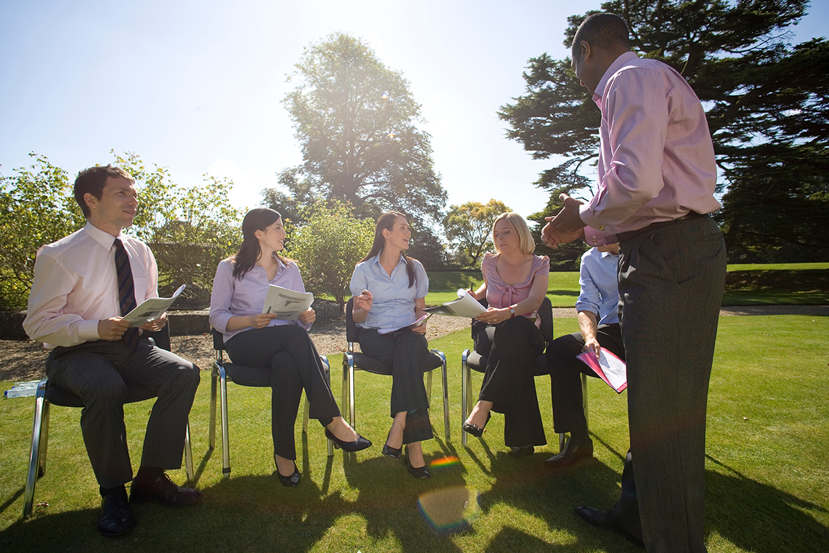 A group of young professionals in business clothes sit on chairs in a grass field while one person is talking to them.