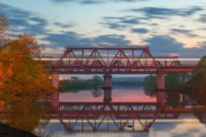 Photo of a rusted red bridge of still water with orange leaved trees on either side.