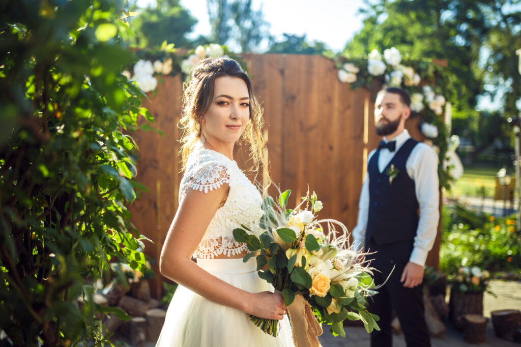 Portrait of a bride in a short sleeved dress holding a bouquet of flowers, looking at the camera. Groom is standing in the background, slightly out of focus.