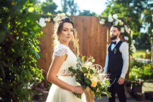 Portrait of a bride in a short sleeved dress holding a bouquet of flowers, looking at the camera. Groom is standing in the background, slightly out of focus.