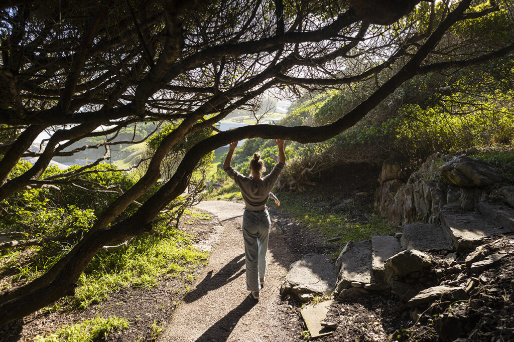 Photo of a woman walking on a path in a wooded area, reaching up to touch a tree above her.